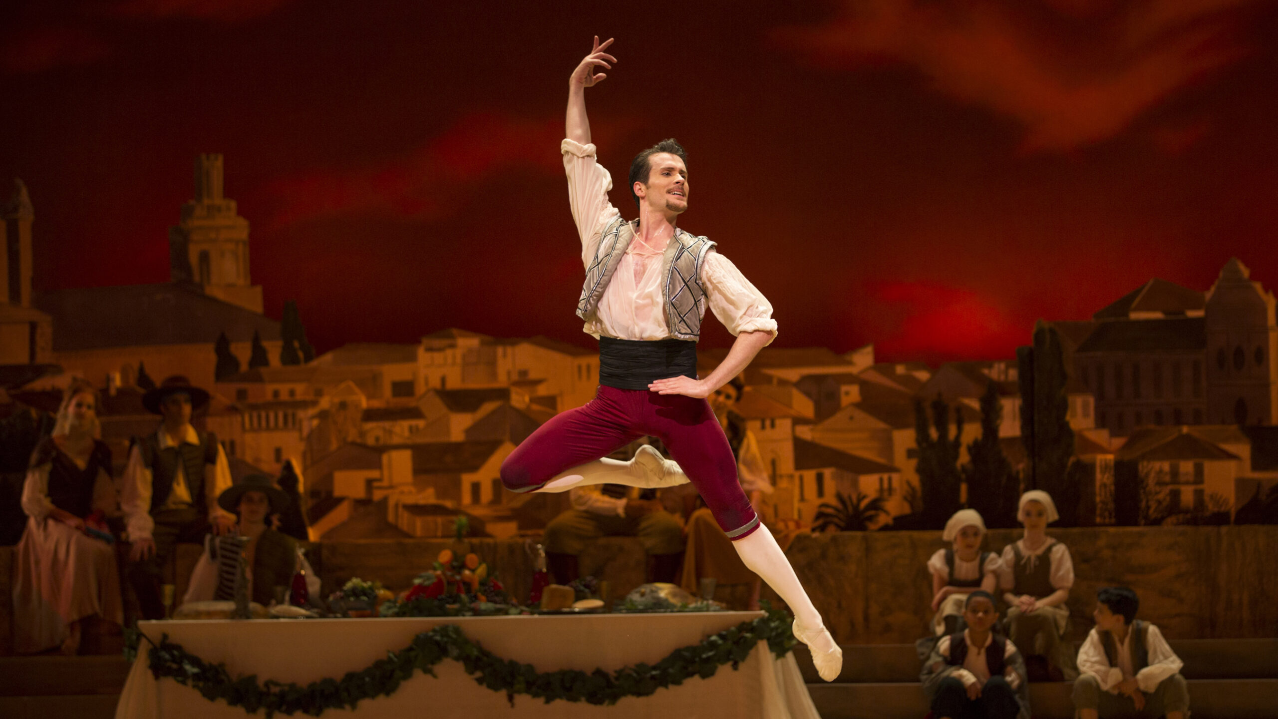 Seth Orza performs in a tavern scene from the ballet Don Quizote. He jumps straight up, his right leg in passé back, and holds his right arm up and places his left hand on his left hip. He looks out over his left shoulder and smiles. Orza wears a white peasant blouse, short gray sleeveless vest, black cumberbund, red knickers and white tights and ballet slippers. A long table full of food is in the background, and other dancers sit and mill about. The set backdrop shows a Spanish city.