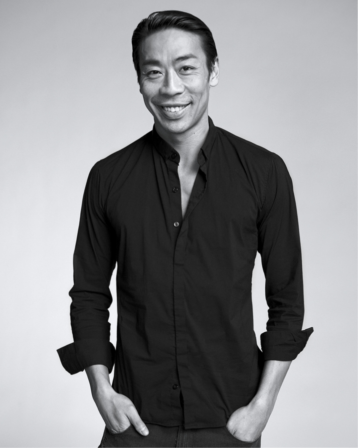 Edwaard Liang, shown hips-up, poses and smiles for a portrait in black and white. He wears a dark button-down shirt and puts his hands in his pants pockets.