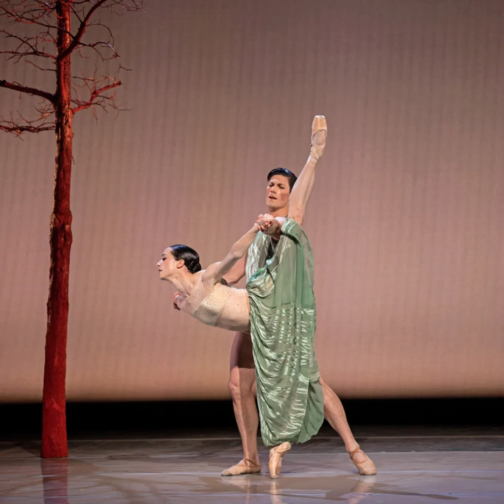 Grace Anne Powers and David Ward perform a pas de deux; she in a penche on pointe and he steadying her from behind, holding each of her hands as she extends her arms behind her.