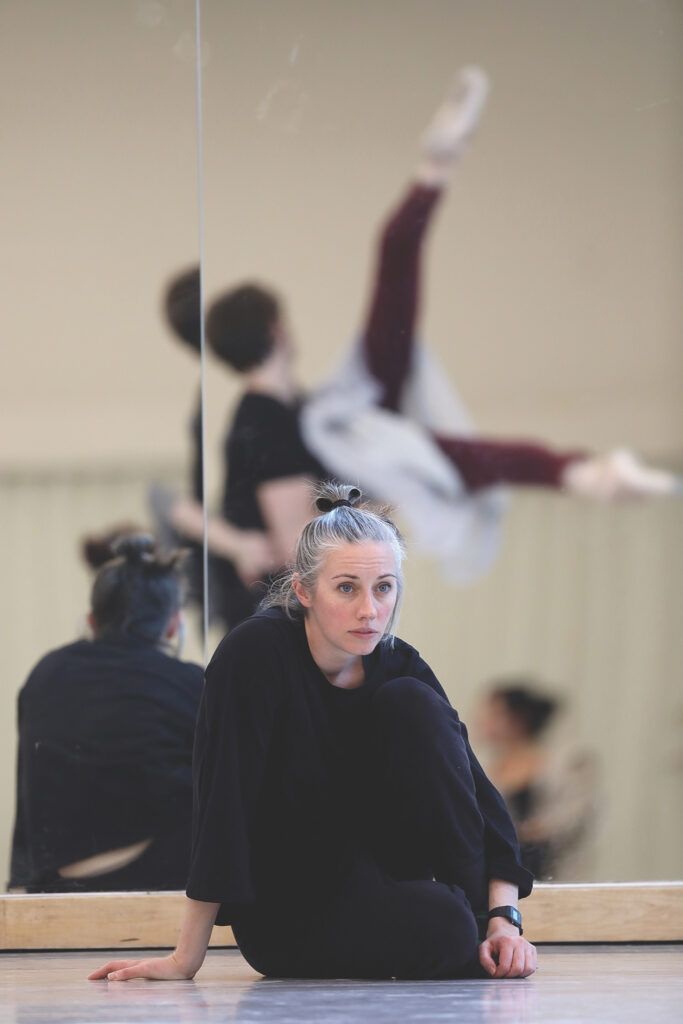 Danielle Rowe, wearing all black, sits on a dance studio floor in front of a mirrored wall and watches a male and female dancer practice a lift.