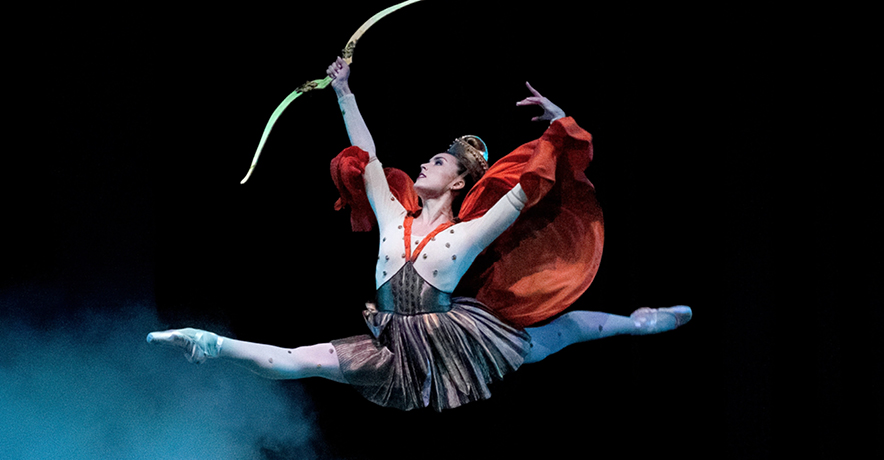 Emily Kikta does a giant leap onstage, surrounding by smoky mist. She holds a bow in her right hand and holds it up triumphantly and lifts her left arm up as well. She wears an off-white and brown dress with a thigh-length pleated skirt, a billowing orange cape, tights, pointe shoes and an elaborate gold headpiece.