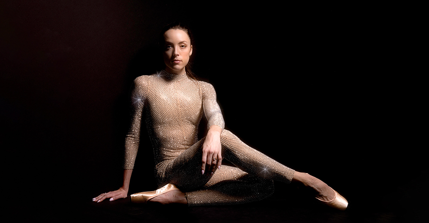 Emily Kikta, wearing a sparkly gold unitard and a new pair of pointe shoes, sits on the ground against a black floor and backdrop. She tucks her left leg under her and crosses her bent right leg over it, resting her left forearm over her thigh. She looks straight ahead with a small, closed-mouth smile.
