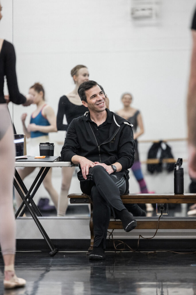 In a ballet studio, Raul Salamanca sits on a wooden bench, his back to a mirrored wall, and crosses his legs. He looks to his left and smiles. In front of him are a group of dancers who stand casually during a rehearsal.