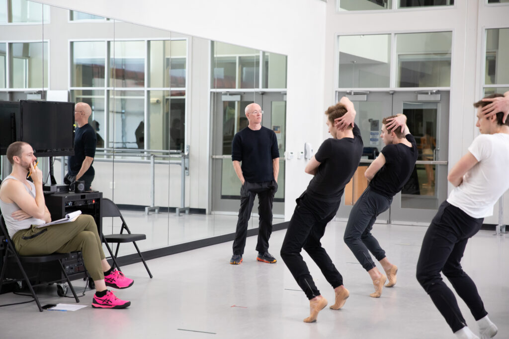 Stephen Mills, wearing a black long-sleeved shirt, black jeans and sneakers, stands at the front of a dance studio and watches a group of male dancers with his hands in his pockets. The three dancers stand with their legs in parallel and push over their feet in demi pointe, bending their knees, thrusting their hips forward, and wrapping their right hand around the back of their heads. Another man, wearing a white shirt, green pants and pink sneakers, sits in a folding chair at the front of the room with a notepad and watches.