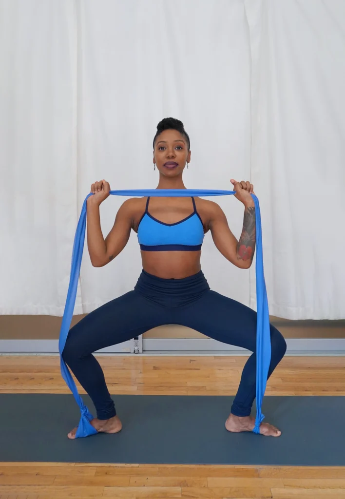 A female dancer in dark blue leggings and a light blue sports bra stands facing the camera and does a grand plié in second position. She holds a long blue resistance band in both hands which she lifts up to her chin. The ends of the band are looped around both feet.