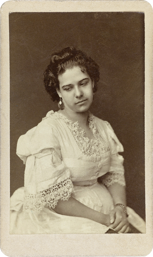 A young 19th century woman in a white dress with pugged sleeves and lace trimmings on the collar and sleeve, sits for a portrait. She crosses her hands in her lap, sits slightly to her left and looks down towards her right shoulder with a closed-mouth smile. Her hair is pulled back at the forehead and piled on top of her head, while the rest of her hair is looslely pulled back into a low bun.