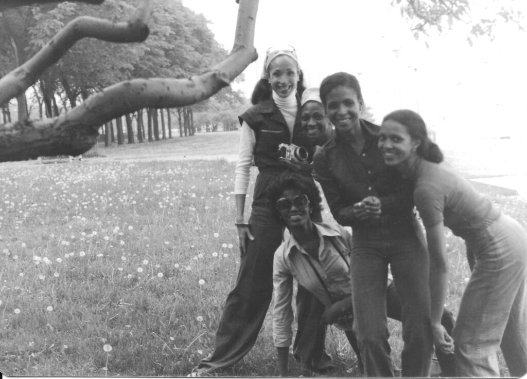 Five Black women wearing jeans and casual shirts stand close together and pose in a field next to a large tree branch.