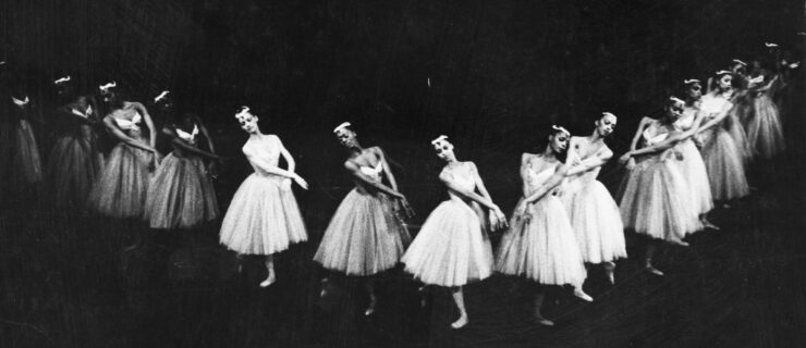 In this black and white photo, a corps de ballet of women stand in a V-shape formation, facing into each other. The dancers stand in tendu croise derriere and cross their ramsa at the wrists in front of them. They wear white knee-length white tutus, white feathered headpieces, tights and pointe shoes.