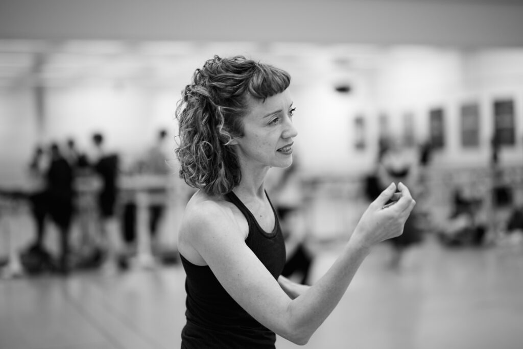 A close-up black and white photo of Cathy Marston leading rehearsal. She speaks and gestures with her right hand.