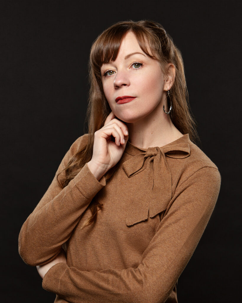 Racheal Nye is shown from the waist up posing in front of a black backdrop. She wears a brown long-sleeved shirt with a bow at the collar, and she rests her right elbow on her left arm and touches her face with her right pointer finger. She looks toward the camera with a closed mouth smile and ears her long brown hair down.