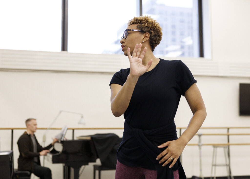 Amy Hall Garner, shown from the hips-up, demonstrates a movement as she leads rehearsal at New York City Ballet. She bends her right arm and rests the back of her right fingertips on the side of her cheek, head turned slightly to the side.