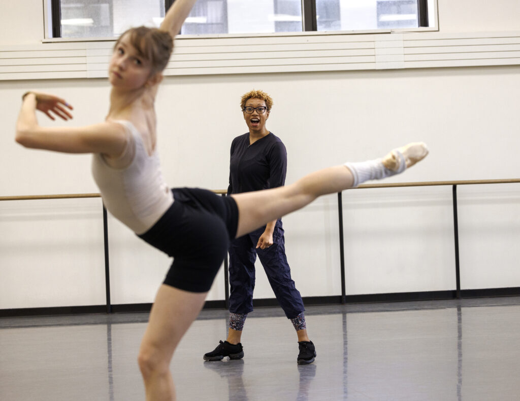 In rehearsal, Emma Von Enck does an arabesque with her supporting leg bent, looking forward toward the front of the studio. Behind her, Amy Hall Garner walks and watches.