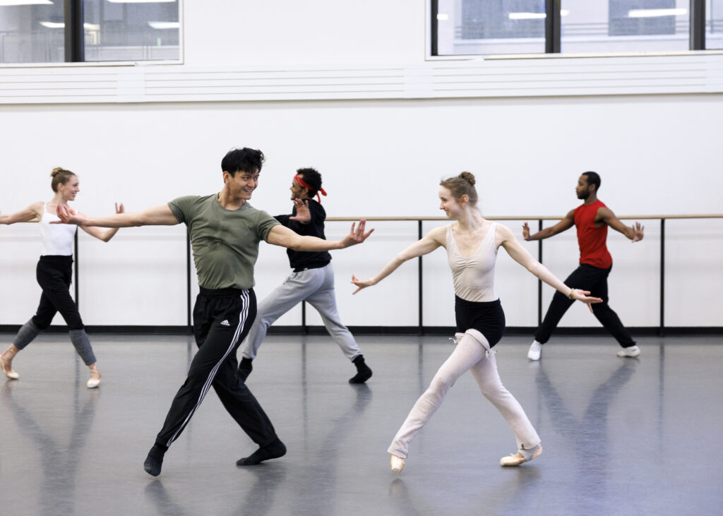 Emma Von Enck and Chun Wai Chan dance together in the foreground of a rehearsal shot, with three other dancers mirroring their movements in the background.
