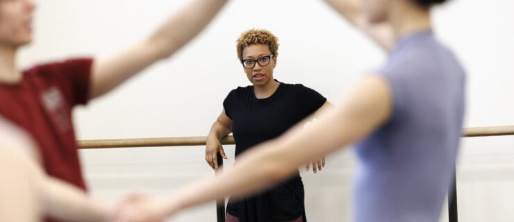 Amy Hall Garner rests her arms on a wall barre in a dance studio as she leads rehearsal. In the foreground, two dancers hold hands and lift their right arms up to frame Garner in the background.
