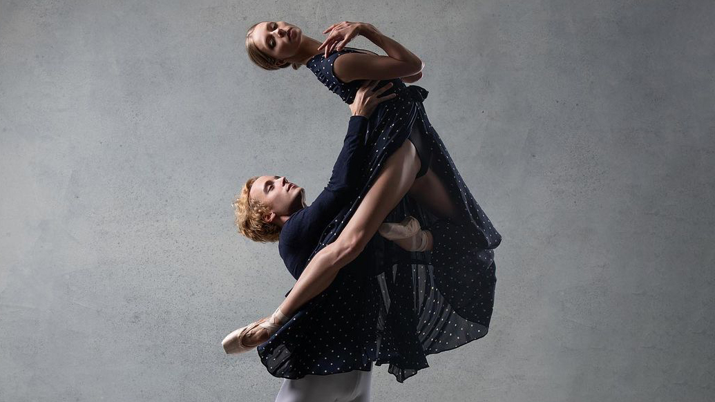 Jonathan Klein holds Emily Hayes up in the air as she arches back, tucking her left leg into passé as she bends her right knee around his ribcage. She holds her hands close to her collarbone and looks out over her right shoulder. Hayes wears a long, blue polka-dotted dress with a large slit up the middle, a dark leotard underneath and pink pointe shoes. Klein wears a dark jacket and white tights.