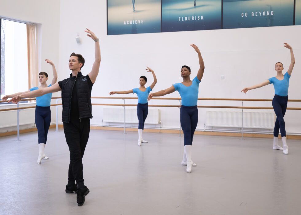 A class of male ballet students in black tights and blue shirts do a tendu combination behind their teacher.