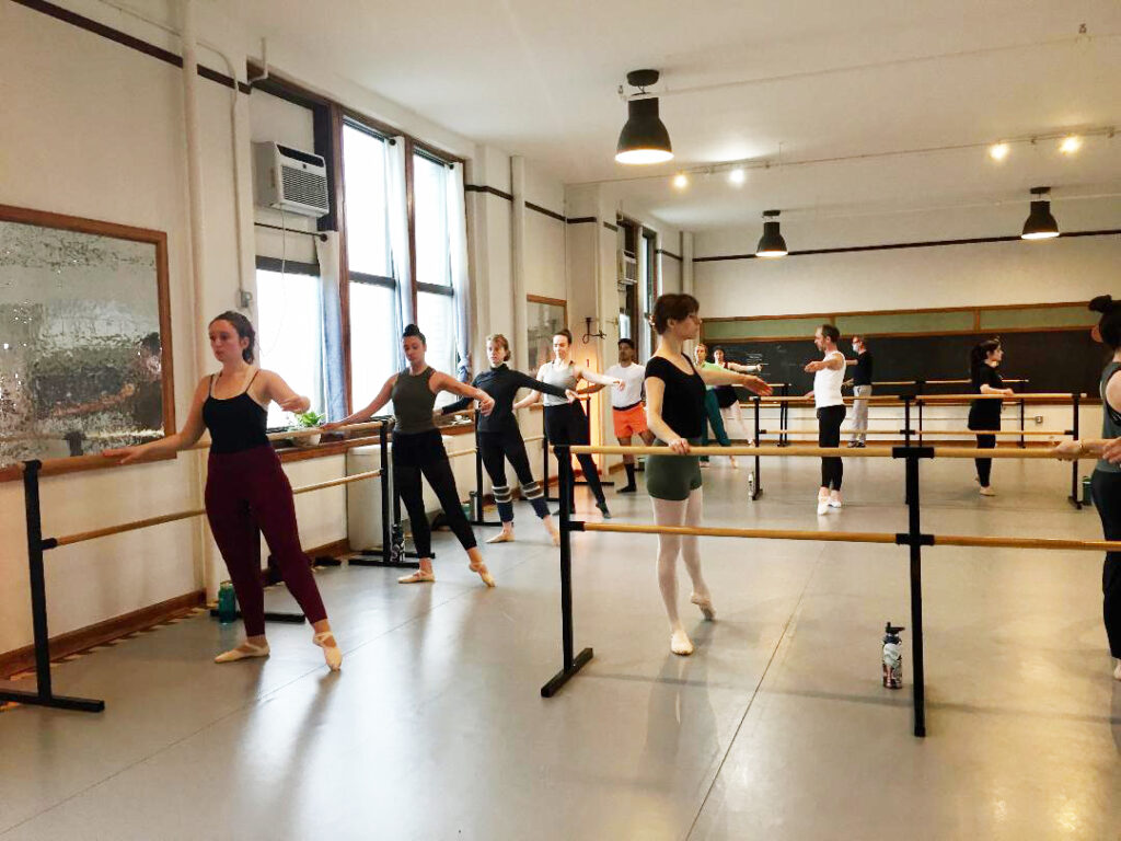 In a large dance studio, a class of adult ballet students perform a tendu exercise at the barre. They extend their right leg and arm in second position and wear various dance outfits.