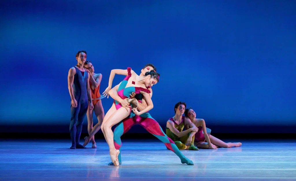 Mikaela Santos, wearing a bright, multi-colored leotard, hinges over her pointe shoes and arches back slightly to her left, looking down and smiling, as a male dancer holds her around the waist. Two sets of couples watch them, with the couple on the left standing and the couple on the right sitting on the ground.