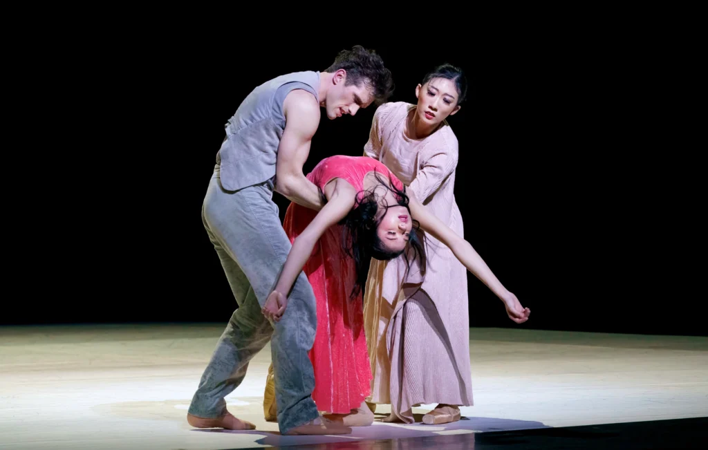Mikaela Santos arches back deeply, her eyes closed and her arms flung above her head, as a male a female dancer catch her around the waist. Santos wears a long pink dress, while the male dancer wears gray pants and sleeveless top and the woman wears a light pink, long-sleeved dress.