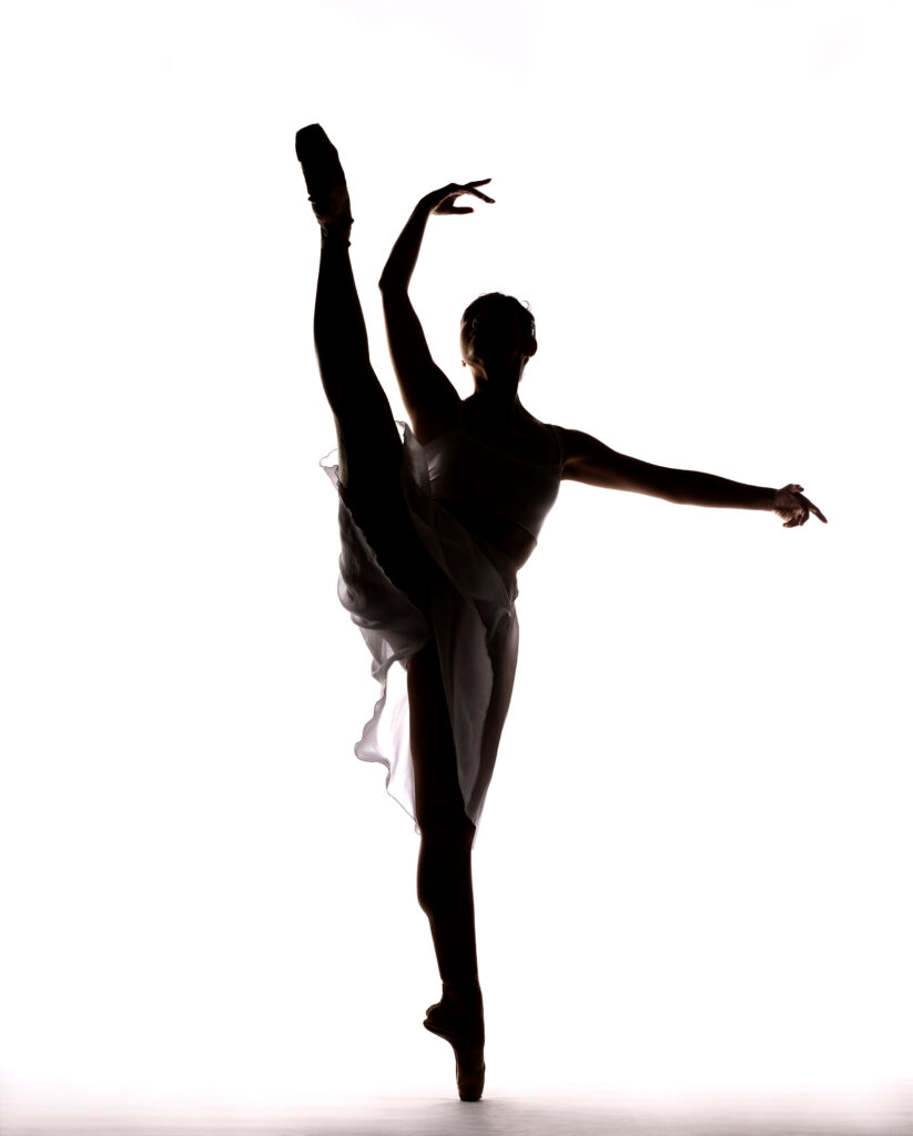 A silhouette of Sarah-Gabrielle Ryan. In front of a white background, Ryan battements her right leg with her arms in high third.