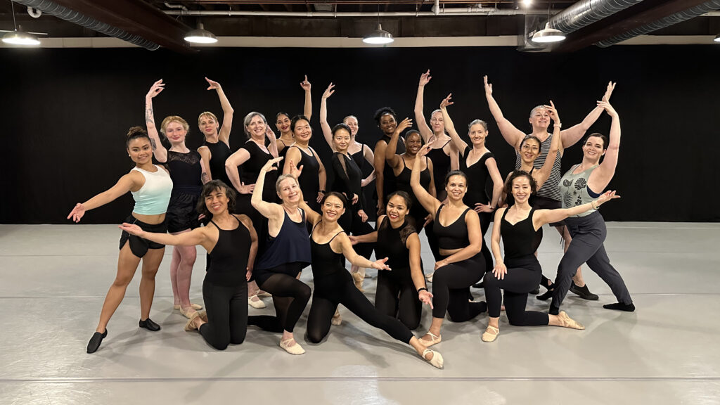 A large group of adult ballet students wearing various dance clothing stand close together and pose for the camera. Six women kneel on the ground while the other dancers stand around them, all with their arms in various ballet poses. They all smile birhgtly for the camera.