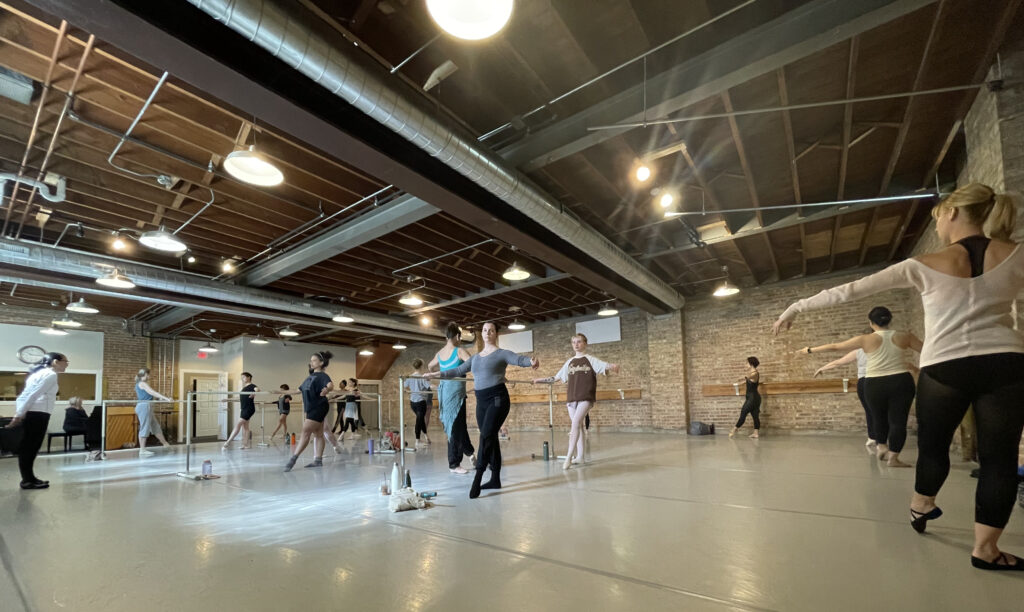 A group of adult ballet students perform a tendu combination at the barre during a dance class. They work in a large dance studio with exposed brick walls and wear various dance clothing.