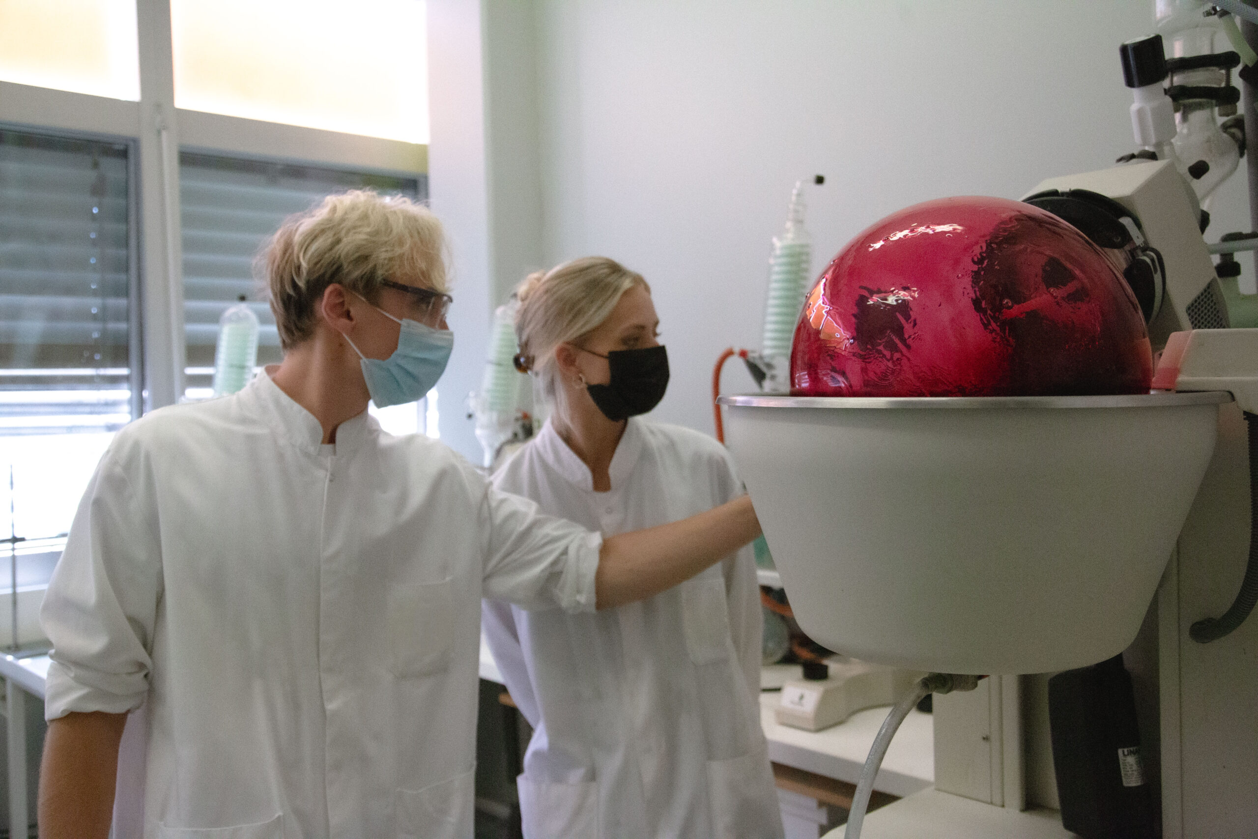 Jonathan Klein and Emily Hayes, both wearing white lab coats and face masks, look at and touch a large white machine in a lab.