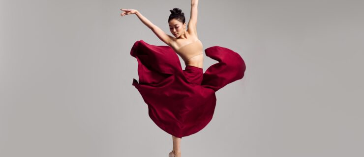 Minori Sakita jumps straight up into the air, her ihps and legs facing her left as she twists her upper body to the right. Her legs are in parallel with pointed feet, and the dancer raises her arms up in a V shape as she looks down towards her right. She wears a tan bandeau top and a long burgundy skirt, which billows up around her as she jumps.