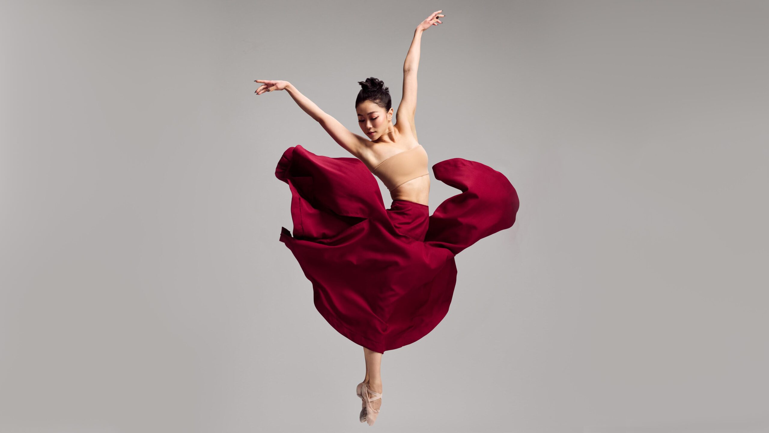 Minori Sakita jumps straight up into the air, her ihps and legs facing her left as she twists her upper body to the right. Her legs are in parallel with pointed feet, and the dancer raises her arms up in a V shape as she looks down towards her right. She wears a tan bandeau top and a long burgundy skirt, which billows up around her as she jumps.