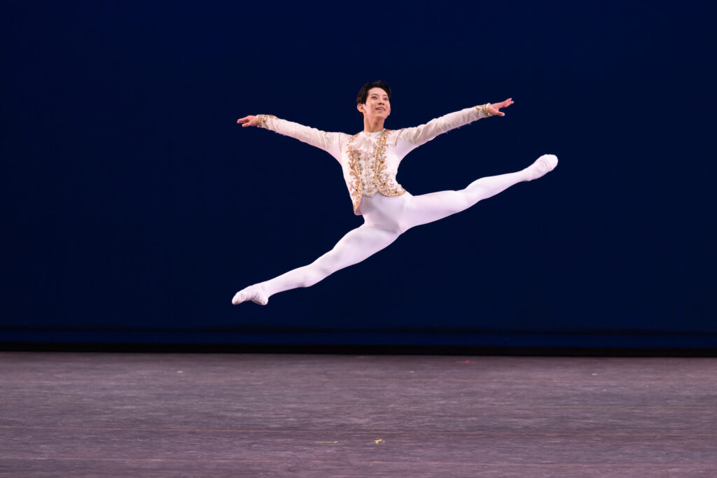 GeonHee Park Performs a sissone onstage towards stage right, his legs split and his arms open wide. He looks out over his left shoulder with a big smile. He wears a white tunic with gold trim, white tights and white ballet slippers.