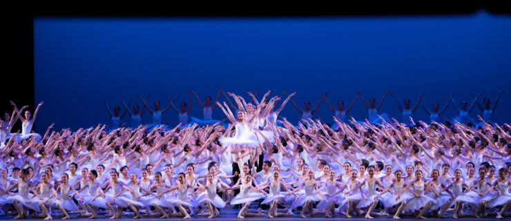 A very large group of student ballet dancers make a tableau, filling the entire stage. The girls wear white leotards, white tutus, pink tights and pink abllet shoes. The boys wear white t-shirts, black tights, white socks and white ballet shoes. The dancers pose in multiple levels, with several lines in the front on their knees, those in the middle in tendu facing the center, and a group of boys in the back and in a line down the center lifting their femal partner into a simple lift.
