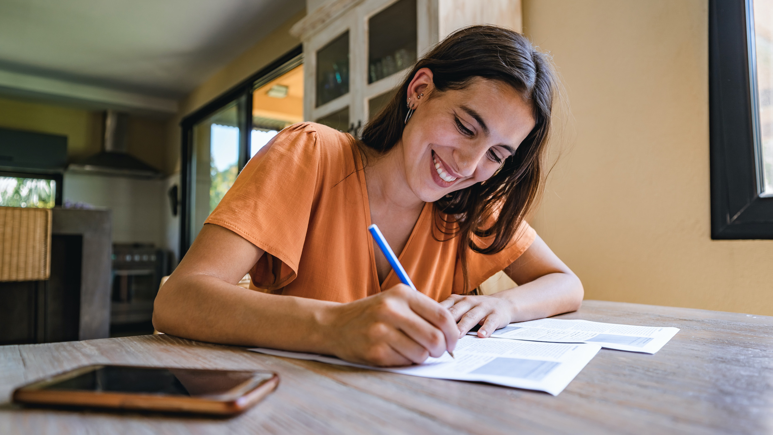 A smiling young woman in an orange shirt sits at a desk and signs papers.