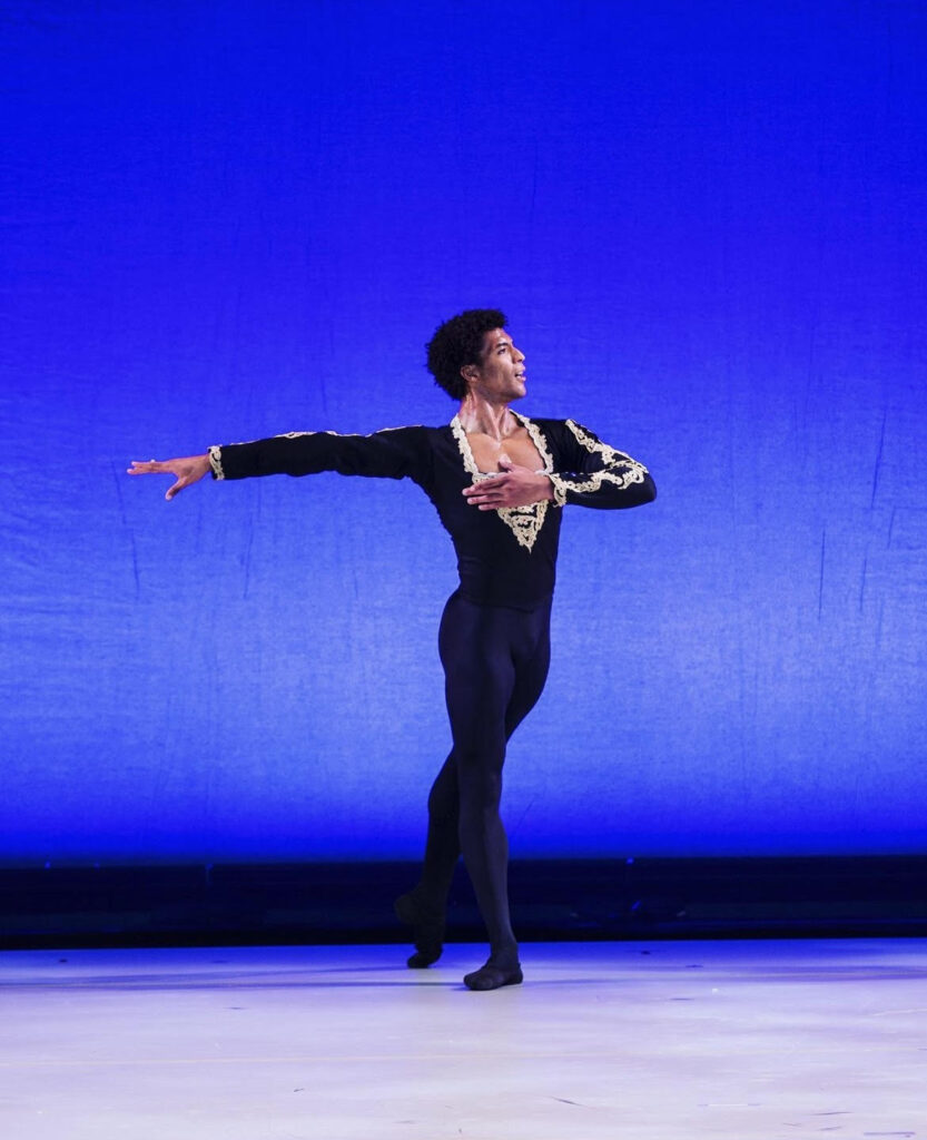 Gian Carlo Perez poses in a tendu derriere onstage, one arm extended and the other folded proudly across his chest.