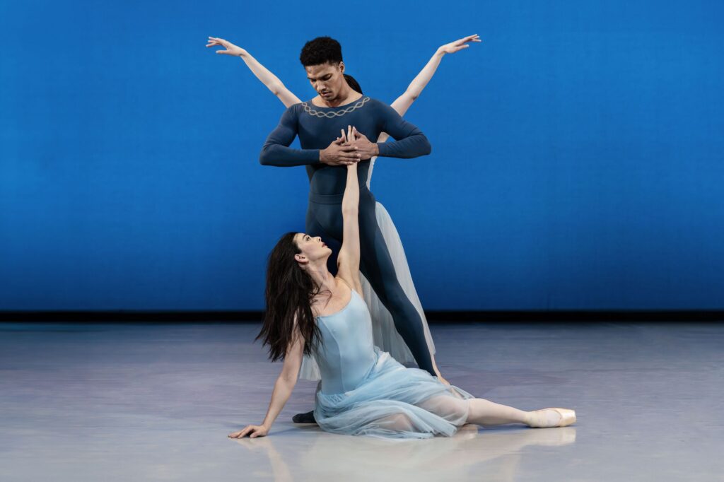 A ballerina in a long blue tutu sits on her right hip on the stage floor, her legs extended to the side. She reaches and looks up to a male dancer wearing a dark blue unitard, who holds her left hand to his heart and looks down at her. Another ballerina stands behind him and reaches her arms out into a high V-shape. The rest of her body is not visible. They dance in front of a blue backdrop.