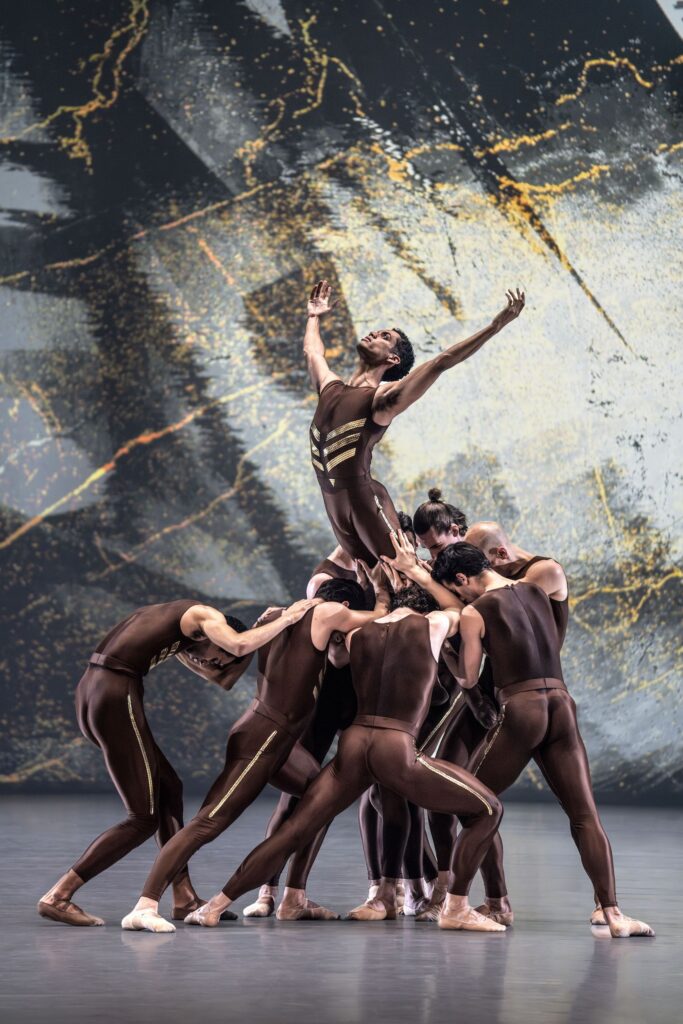 A group of male dancers in brown unitards clump together, heads down, and lift up another male dancer. The lfited dancer arches his back slightly and lifts his arms up, palms in, and looks up toward the upstage corner. They dance in front of a gray backdrop with a mottled yellow design.