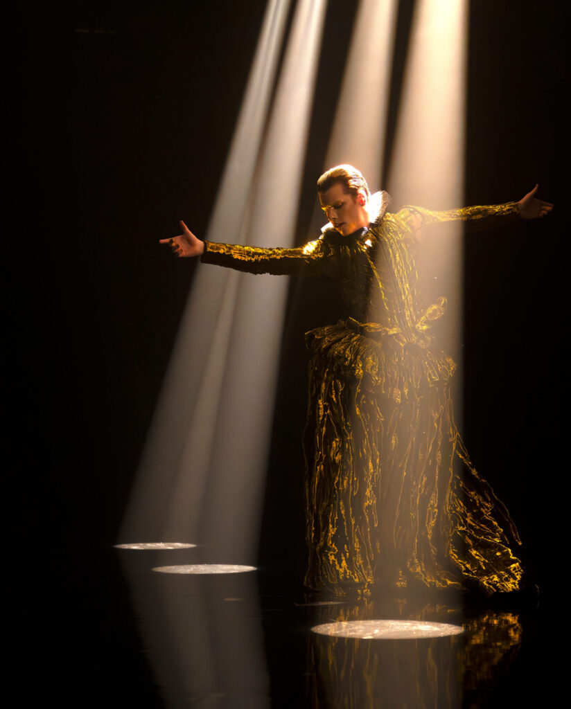Jake Roxander, in a large gold costume with a white ruffle neck, stands between four stark columns of light on a black stage.