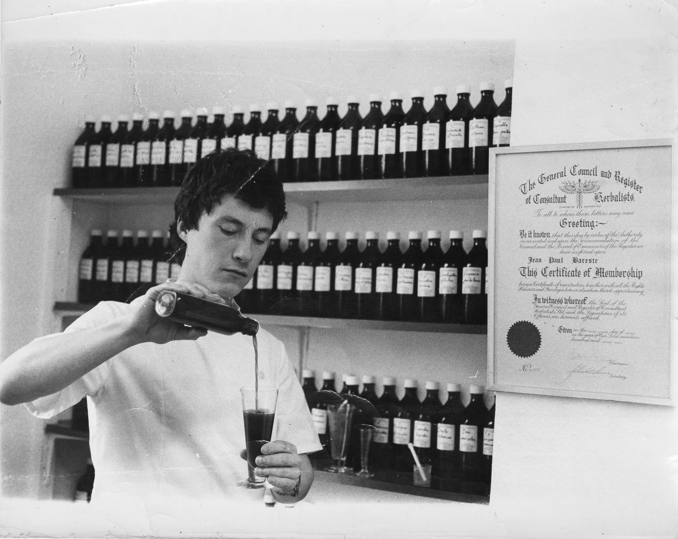 In this black and white photo, Jean-Paul Barestepours a dark liquid out of a bottle and into a glass. He wears a white lab coat and stands in front of three shelves of bottled liquids, and a diploma hangs on the wall to his right.