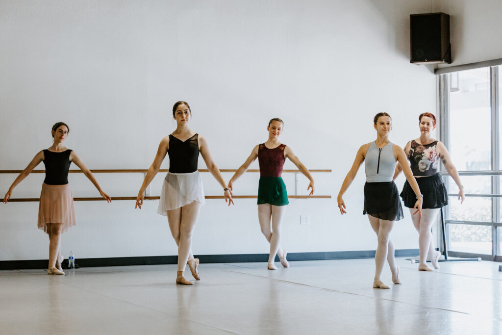 A group of five adult ballet students stand in b plus with their right foot back and their arms in demi seconde. They all wear leotards, pink tights, ballet slippers and short ballet skirts in various colors. They stand in a staggered line in a spacious dance studio, with three dancers in back and two in front.