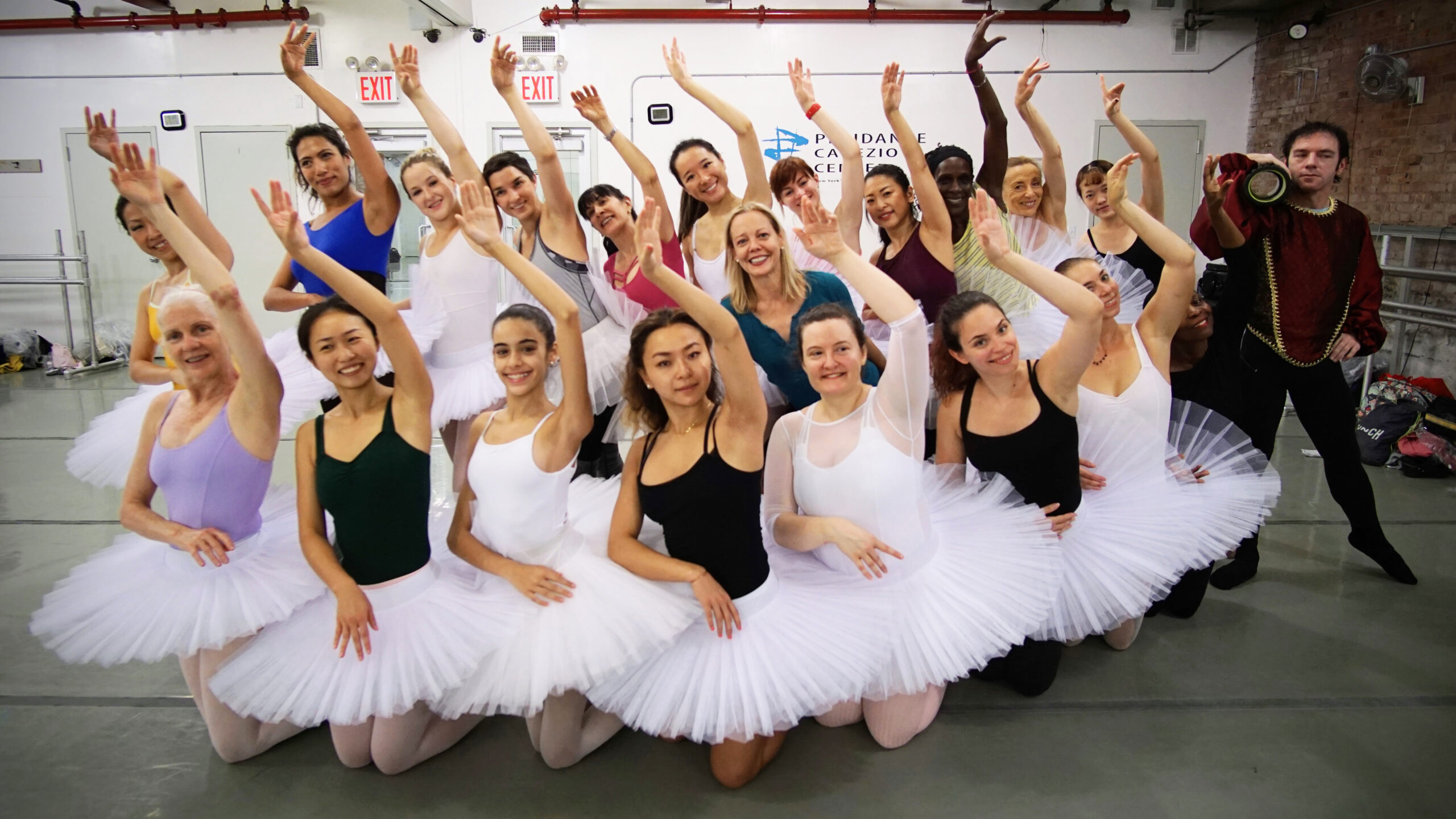 A large group of adult ballet dancers wearing leotards in various colors and white practice tutus pose closely together with their left arm raised next to their ear, the elbow slightly bent and the palm facing out. Their right arms are bent at the waist, crossing in front of their bodies. Seven of the dancers kneel in the front row, while the others stand. An adult male dancer stands at the far left of the group, posing in a tendu with his left hand on his hip as he holds a foam roller on his right shoulder.