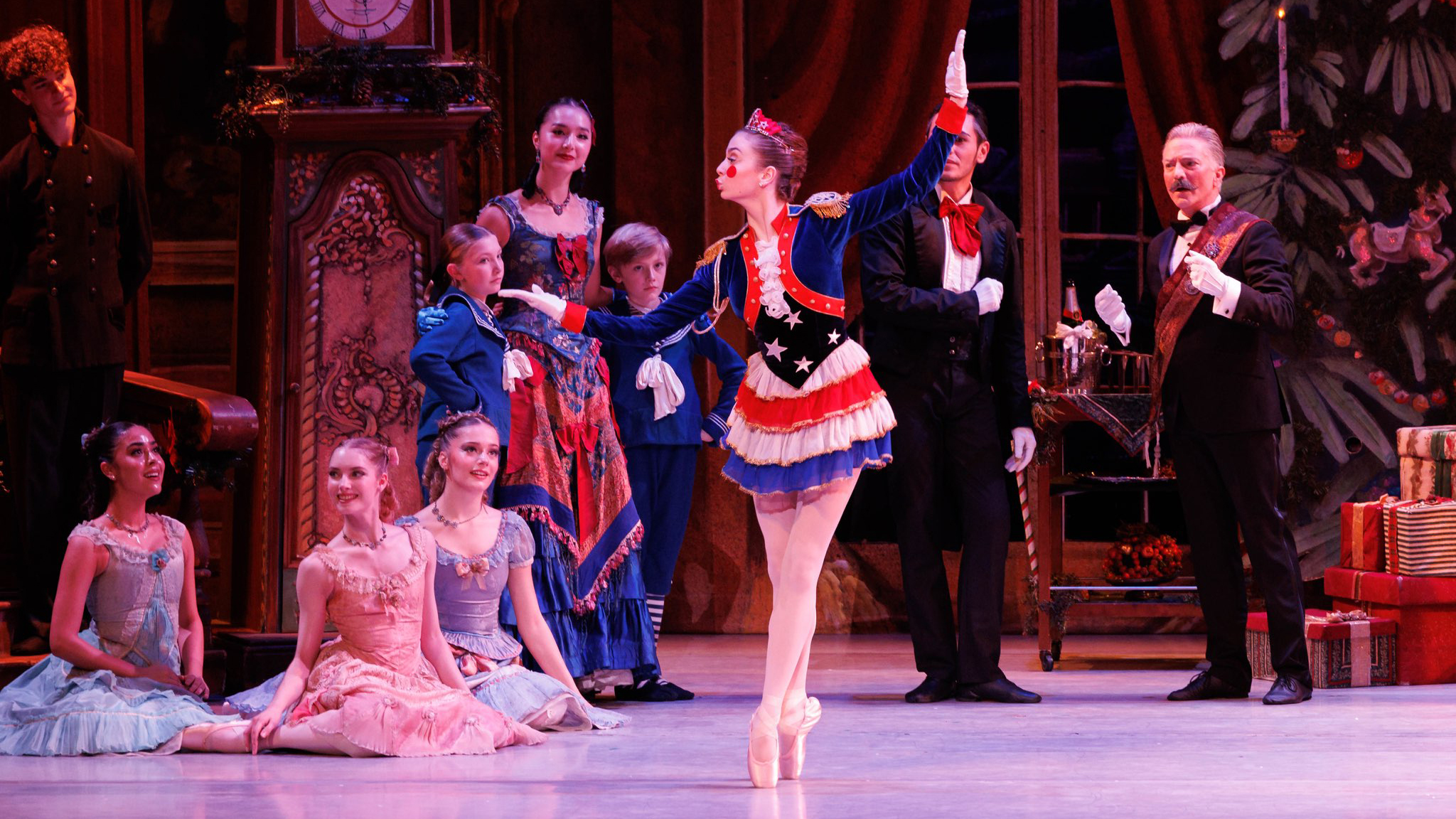 Abigail Brent stands onstage in sus-sous onstage. She holds her arms out to the side, bent at the elbows like a mechanical doll and kisses the air. She wears a red white and blue velvet jacket and short skirt, with a star-spangled cumberband, pink tights, pointe shoes and red crown. She also has red felt circles glued onto her cheeks. Young dancers sit on the floor and watch her dance.