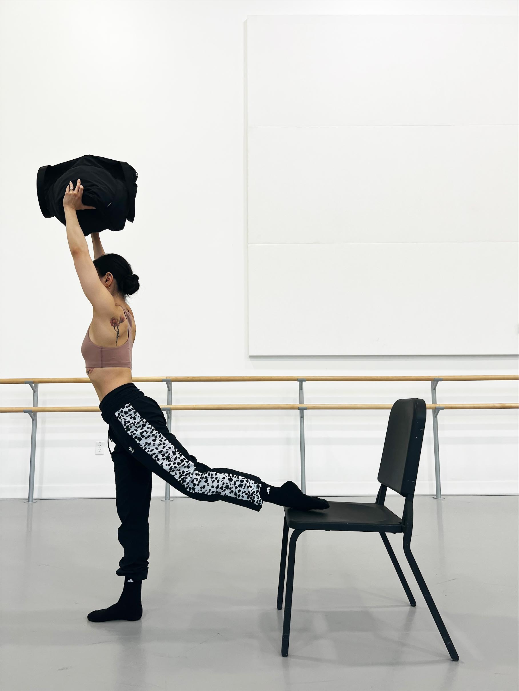 Minori Sakita, wearing a brown sports bra, black workout pants and black socks, stands in front of a black chair and places the top of her left foot on the seat. Her left leg is slightly bent behind her. She holds up a black bag above her head.