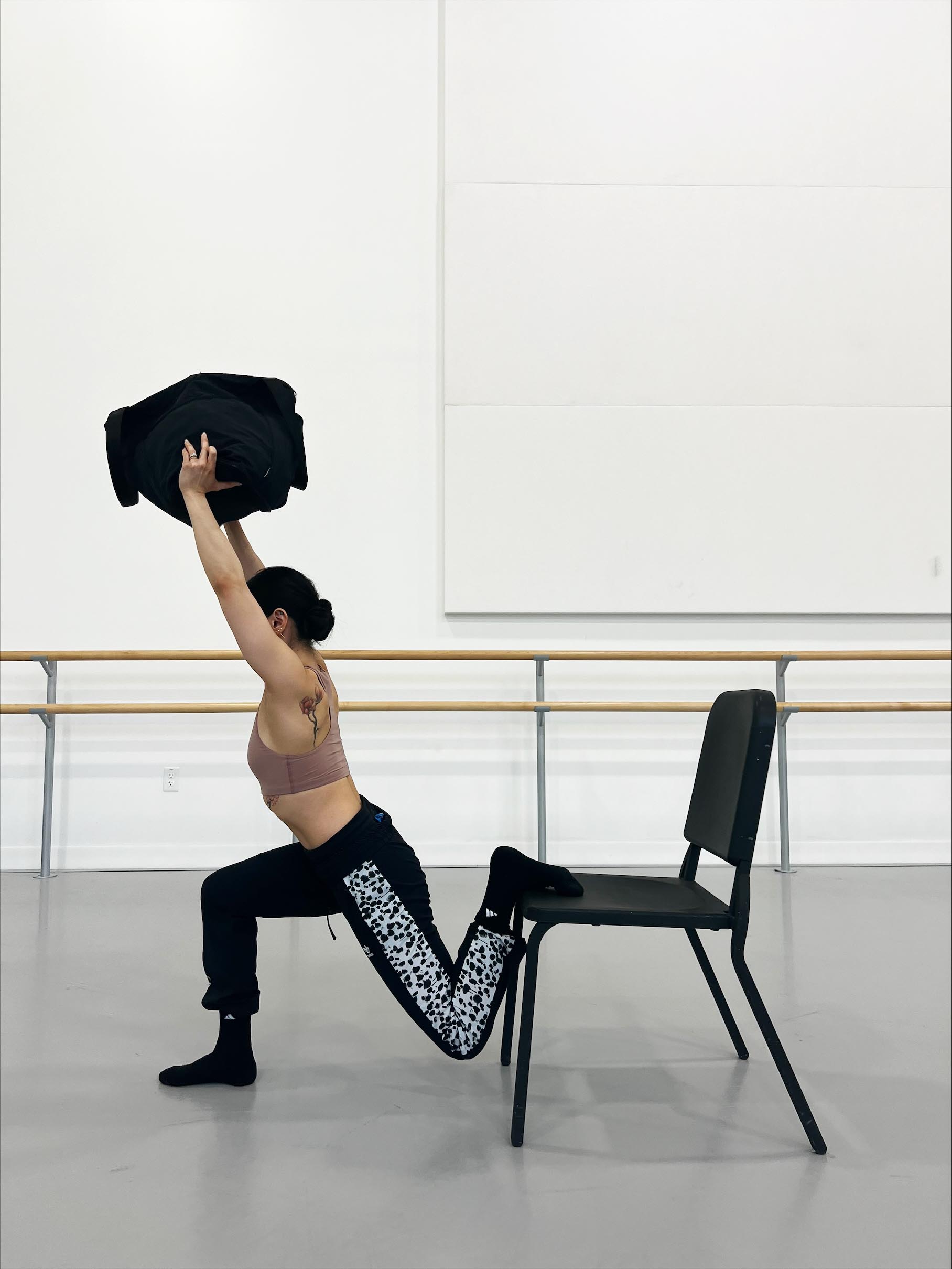 Minori Sakita, wearing a brown sports bra, black workout pants and black socks, stands in front of a black chair and places the top of her left foot on the seat. She squats down on her right leg, wit her left leg bent behind her. She holds up a black bag above her head.