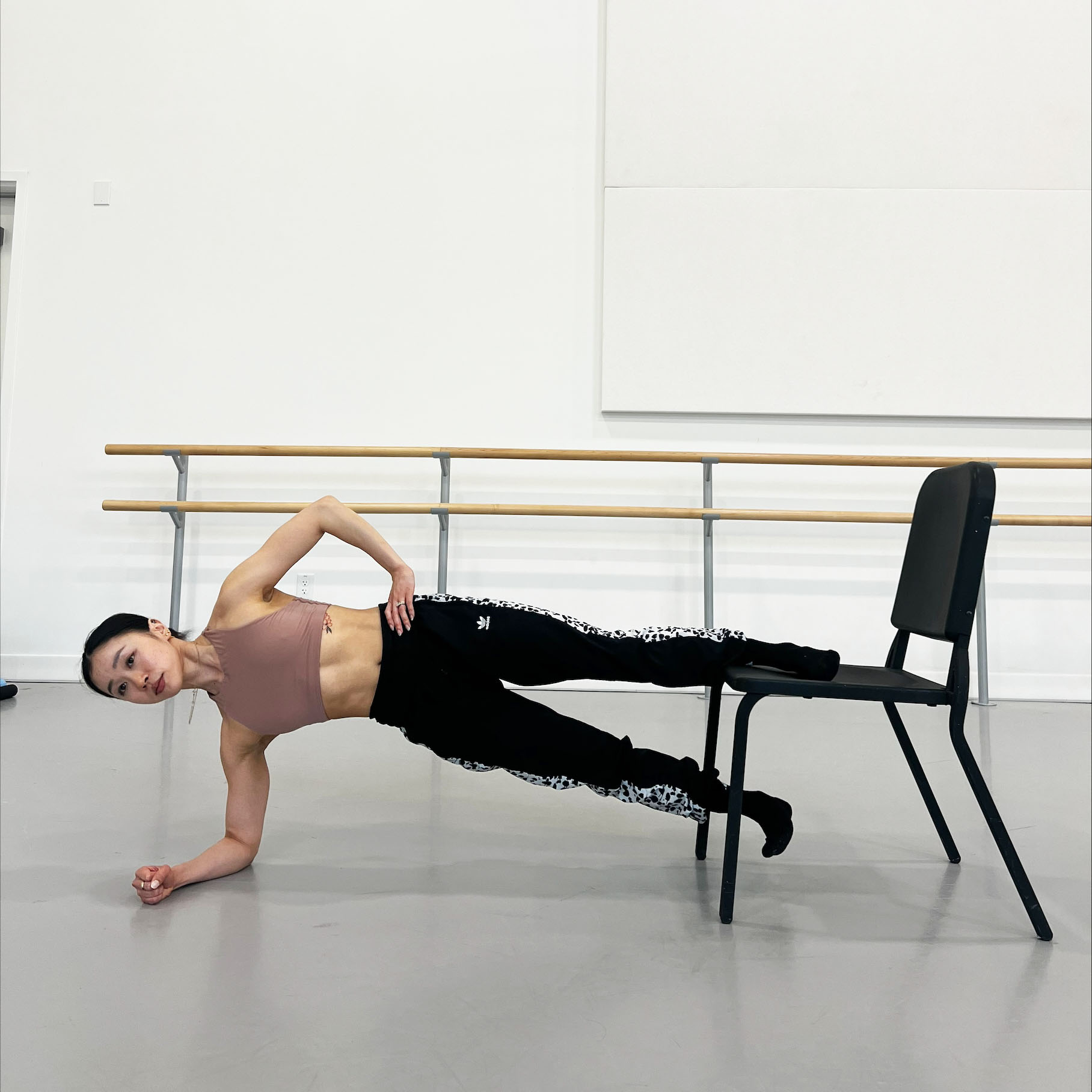 Minori Sakita, wearing a brown sports bra, black workout pants and black socks, balances in a plank on her right forearm with her left foot on the seat of a black chair. She lifts her straight right leg up towards the bottom of the chair's seat. flexing her foot.