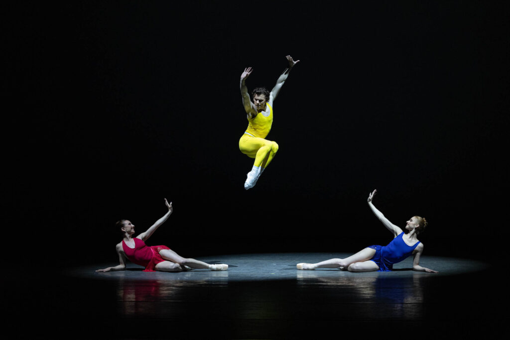 On a drak stage and in a bright spotlight, Jake Roxander, in a bright yell unitard, jumps straight up, tucking his legs underneath him as he lifts both arms up over his head. Isabella Boylston and Catherine Hurlin lie on the ground on either side of him, propped up on their elbow and lifting their other arm up witha. flexed wrist. Boylston is costumes in a red dress and Hurlin in a bright blue dress. Both wear pointe shoes.