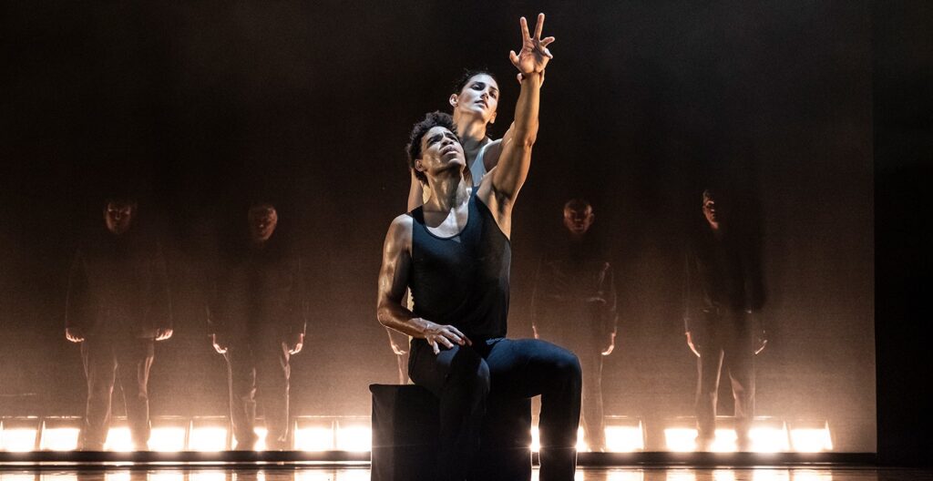 Laura Rodriguez stands behind Carlos Acosta, who sits on a black box, and matches the line of his arm as they reach up and out toward the audience.