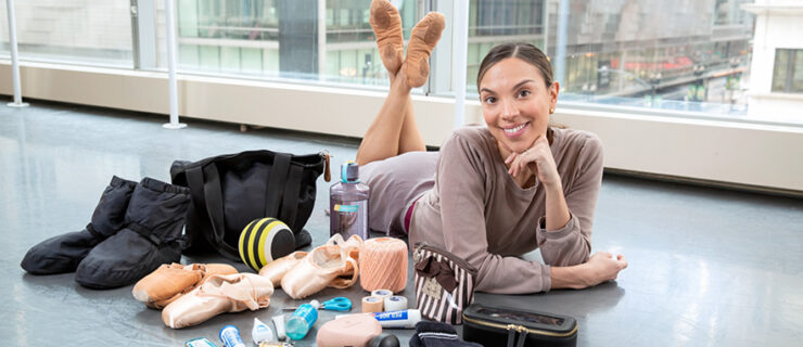 Anais Bueno, wearing a mauve long-sleeved shirt and ballet skirt, lays on her stomach on a dance studio floor and props herself up on her elbows, her left hand under her chin. She lifts her legs behind her, crossing her feet, and smiles for the camera. To her right are the contents of her black dance bag, such as shoes, massage tools, and makeup bags, spread out onto the floor.