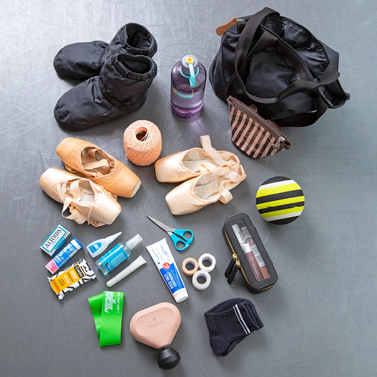 An array of dance bag items are neatly arranged on a dance studio floor and shown from above. They include dance shoes, booties, 2 zip-up pouches, a roll of pink crochet thread, 3 rolls of sports tape, various massage tools, ointment, scissors, mints and snacks.