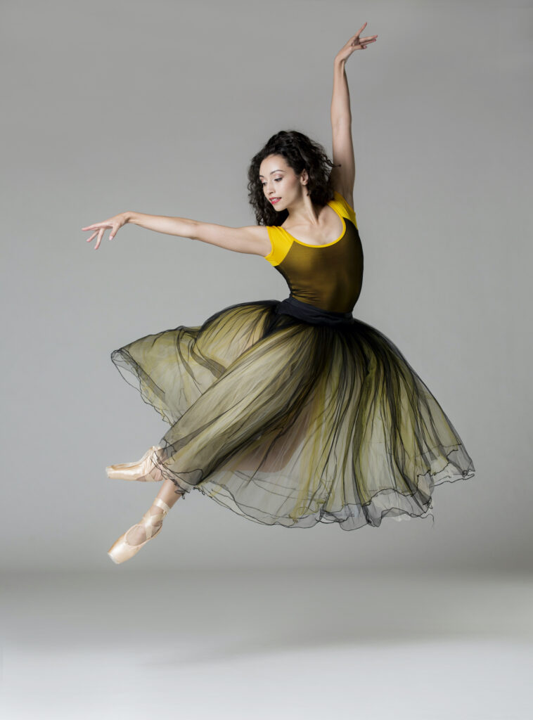 Sarah Gabrielle Ryan does a jeté sauté, arching her body slightly to the right. She holds her left arm out and her right arm out to the side and looks out over her right hand. She wears a long tutu with black and yellow tulle, a yellow and black mesh leotard, and pointes shoes, and she dances in front of a gray backdrop.