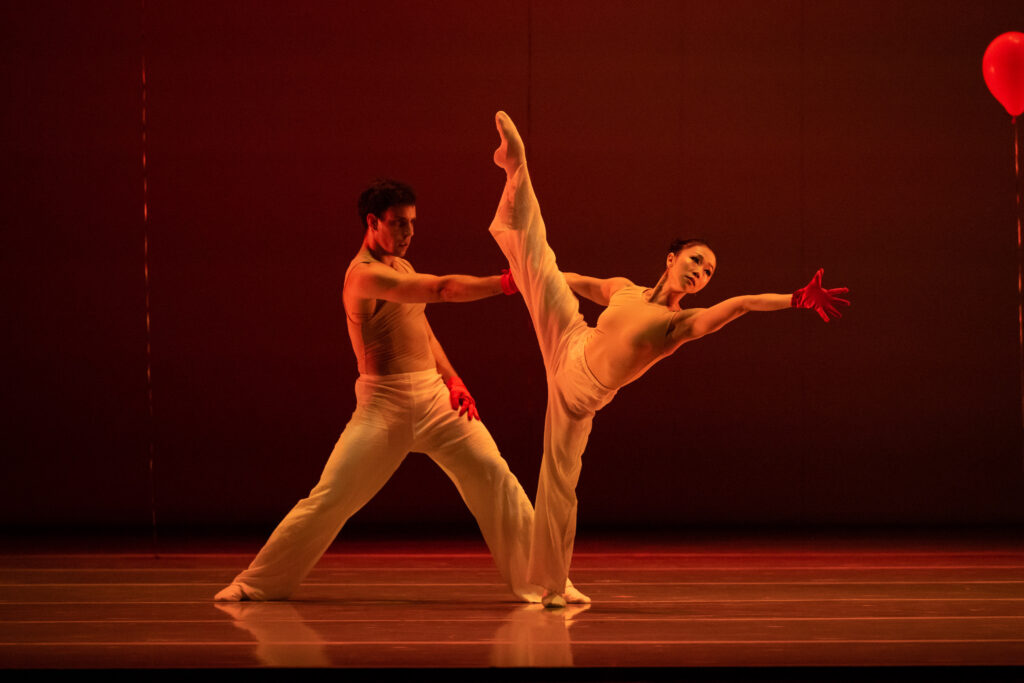 During a dance performance, Taylor Carrasco takes a wide stance and reaches out his right arm, holding the hand of Minori Sakita as she pulls away from him and lifts her right leg high to the side. She reaches out and away with her left hand, looking off into the distance in that direction. Both dancers wear red gloves, offwhite pants and tan-colored tops.