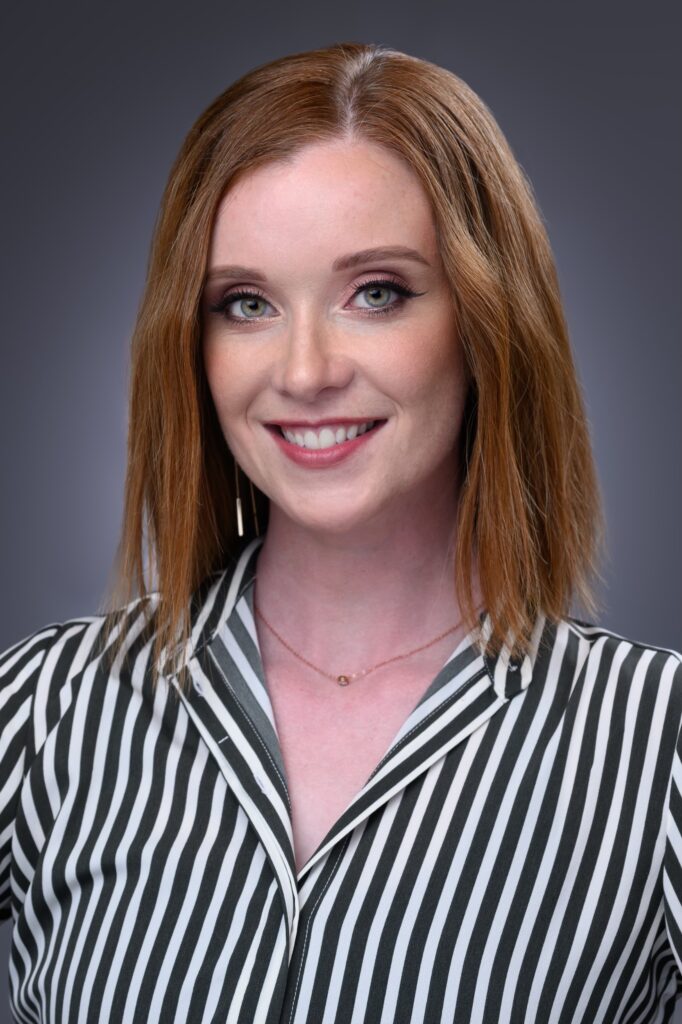 Tracy Jones is shown from the chest up. She wears a button-down shirt with black and white vertical stripes and a delicate necklace. She wears her shoulder length red hair down and smiles directly at the camera.